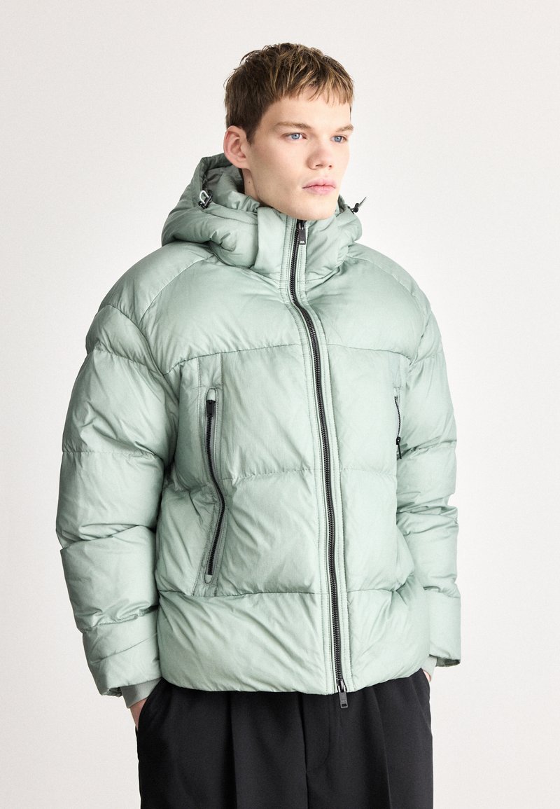 Απαλό πράσινο μπουφάν puffer με κουκούλα, διαθέτει εμπρόσθιο φερμουάρ, τσέπες με φερμουάρ στα πλάγια και καπιτονέ υφή.