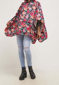 Kwiatowy ponczo w ciemnym materiale z efektownymi różowymi i pomarańczowymi kwiatami, luźny krój, kaptur i otwarte boki. Noszone na jasnoniebieskich, przetartych dżinsach.