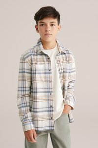 Rutigt flanellskjorta i vitt, blått och brunt. Mjuk textur med knappar och bröstficka. Bärs över en ljus t-shirt och gröna byxor.