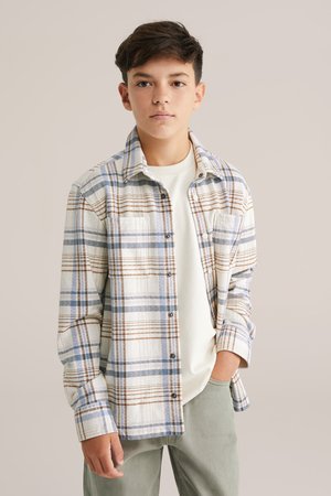 Rutigt flanellskjorta i vitt, blått och brunt. Mjuk textur med knappar och bröstficka. Bärs över en ljus t-shirt och gröna byxor.