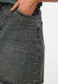 Jupe en denim noire, présentant une texture douce, avec des clous en argent disposés de manière éparse le long de la poche latérale pour un détail supplémentaire.