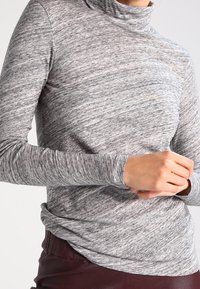 Frau trägt einen enganliegenden graumarbigen Langarm-Turtleneck und dunkelburgunderfarbene Hose, mit gefalteten Händen auf der Taille.