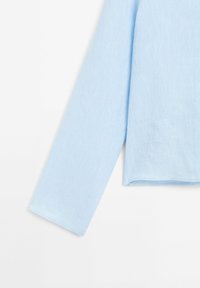 Manche longue d'une chemise bleu clair et partie du torse sur fond blanc, montrant la texture du tissu et les détails des coutures.