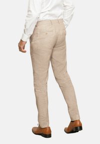 Pantalones de lino beige con corte slim, que cuentan con bolsillos traseros y un botón, combinados con zapatos de cuero marrón.