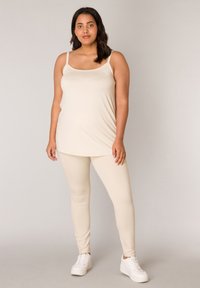 Beige tanktop met dunne bandjes en een losse pasvorm, gecombineerd met bijpassende leggings. Gemêleerde stof, gladde afwerking. Draagt met witte sneakers.