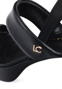 Sandalia de plataforma de cuero negro con diseño curvado, que cuenta con una correa elegante y un acento en color dorado del logo en el costado. Textura suave.