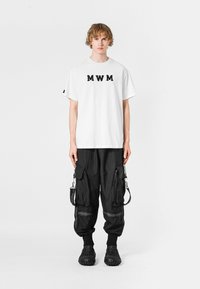 Camiseta blanca de mangas cortas con letras "MWM" en negro, combinada con pantalones cargo negros que presentan múltiples bolsillos y un material texturizado. Calzado negro.
