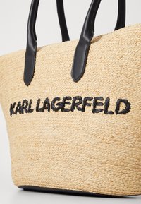 Torbica od pletene slame s crnim kožnim ručkama, sa bordurom "KARL LAGERFELD" izvezenom crnom niti na prednjoj strani.