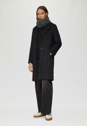 Schwarzer Wollmantel mit Reverskragen, zwei Knöpfen, Seitentaschen; grauer Strickschal, weit geschnittene dunkle Hosen, beigefarbene Schuhe mit schwarzen Akzenten.