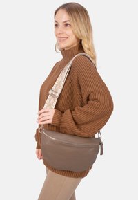 Braune Leder-Umhängetasche mit glatter Textur, geschwungenem Design und silberner Reißverschlussbeschlägen; verfügt über einen gemusterten verstellbaren Tragegurt.