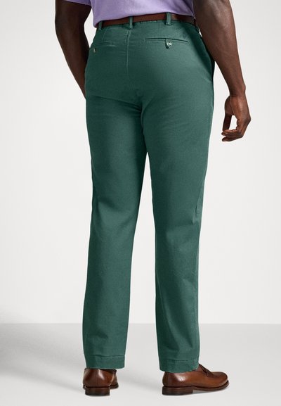 Πράσινο βαμβακερό παντελόνι με ίσια γραμμή, με τσέπες στο πίσω μέρος και θηλιές για ζώνη. Συνδυασμένο με καφέ δερμάτινα παπούτσια.