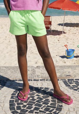 Personne portant un short vert citron, un haut rose et des tongs roses, debout sur un pavement en mosaïque près d'une plage de sable avec un seau et un parasol.