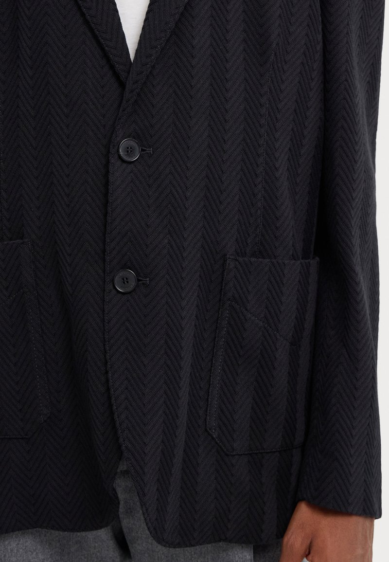 Blazer noir à motif chevrons, doté d'un col cranté, de deux boutons et de deux poches avant avec une finition en tissu texturé.