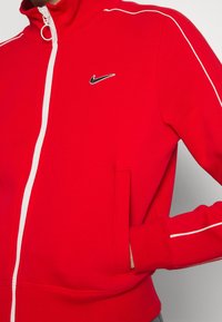 Röd zip-up jacka gjord av mjukt material, med en vit dragkedja och foder, ribbade ärmslut och en liten svart Nike-logotyp på bröstet.