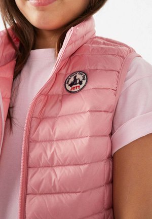 Jongere draagt een lichtroze gewatteerd vest over een lichtroze T-shirt, met een ronde "JOTT" logo patch op de borst van het vest.