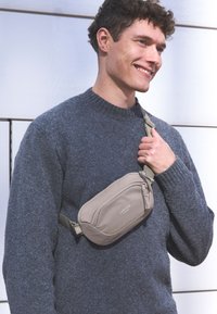 Borsa a vita beige con tasca con zip, indossata trasversalmente sul petto. Il tessuto è liscio e presenta un design minimalista. Il modello indossa un maglione scuro e strutturato.