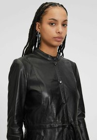 Giacca in pelle nera con colletto alto, chiusura con bottoni e vita regolabile con cordino per una vestibilità personalizzabile, texture liscia con una leggera lucentezza.