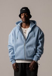 Himmelblaues Zip-Hoodie mit einer vorderen Kängurutasche, Kapuze mit Kordelzug und gerippten Bündchen, getragen über einem weißen Shirt und kombiniert mit schwarzen Shorts.