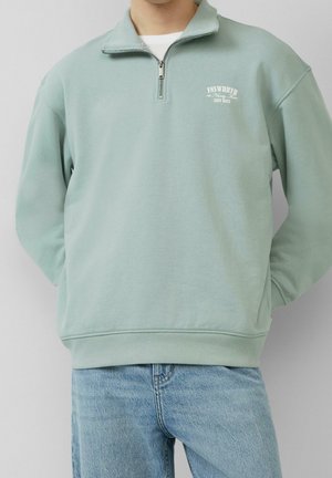 Homme portant un sweat-shirt zippé vert clair avec un petit texte blanc sur la poitrine, associé à un jean bleu clair sur un fond uni.