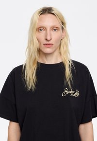 Čierne bavlnené oversized tričko s vyšívaným zlatým logom v oblasti ľavého hrudníka. Mäkká textúra s uvoľneným strihom a krátkymi rukávmi.