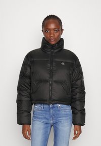 Svart puffjakke med høy krage, frontglidelås, kviltet tekstur og CK-logo på brystet, sammen med lyseblå jeans.