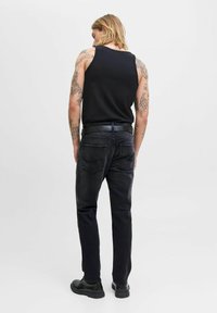 Joven con cabello hasta los hombros y tatuajes en los brazos, vistiendo una camiseta sin mangas negra, jeans oscuros, cinturón negro y zapatos negros, de pie con la espalda hacia la cámara.
