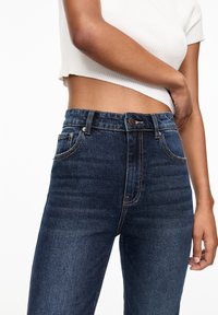 Jeans in denim blu scuro con vita alta, dotati di cinque tasche, cuciture a punto, e chiusura con bottone, abbinati a un top corto bianco a costine.