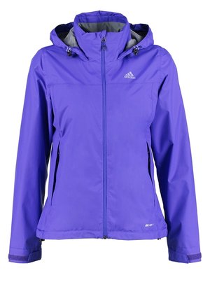 Persona con chaqueta y pantalones cortos púrpuras, camiseta blanca gráfica y botas de senderismo negras caminando hacia adelante sobre un fondo sencillo.