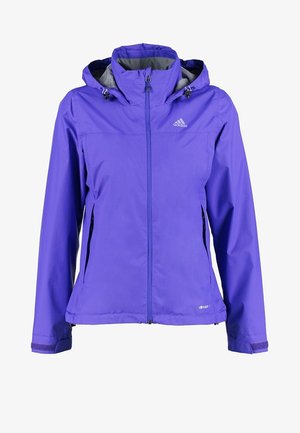 Persona con chaqueta y pantalones cortos púrpuras, camiseta blanca gráfica y botas de senderismo negras caminando hacia adelante sobre un fondo sencillo.