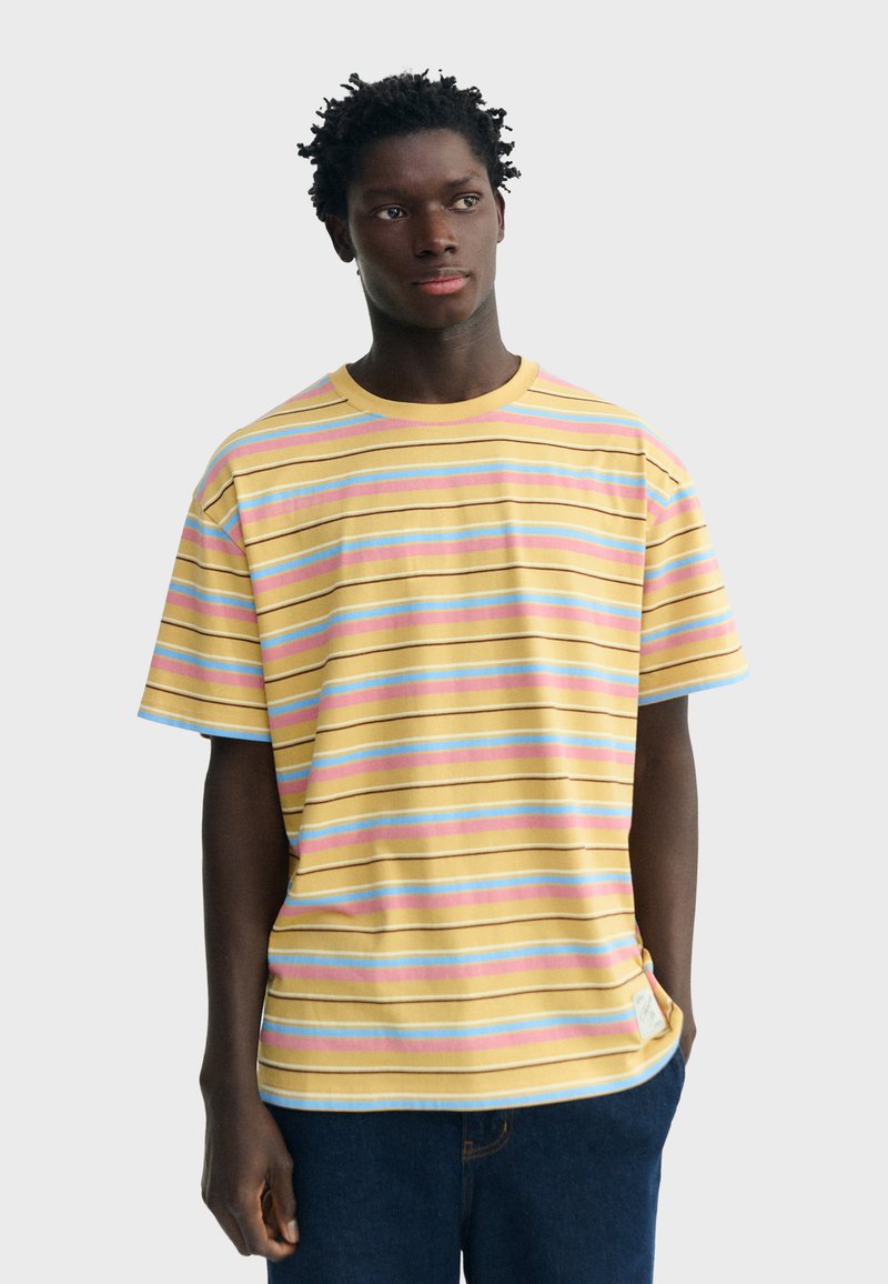 Jeune homme portant un T-shirt jaune à rayures avec des lignes pastel bleu, rose et blanc, debout les mains le long du corps.