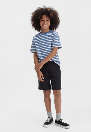 Χαμογελαστό παιδί με σγουρά μαλλιά, φοράει μπλε και λευκό ριγέ μπλουζάκι, μαύρο σορτς, γκρι κάλτσες και μαύρα αθλητικά, στέκεται με τα χέρια σταυρωμένα.