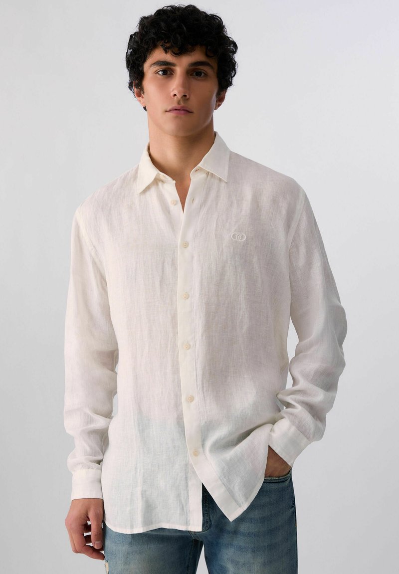 Jeune homme aux cheveux bouclés portant une chemise blanche en lin à manches longues et un jean bleu, debout avec une main dans la poche.
