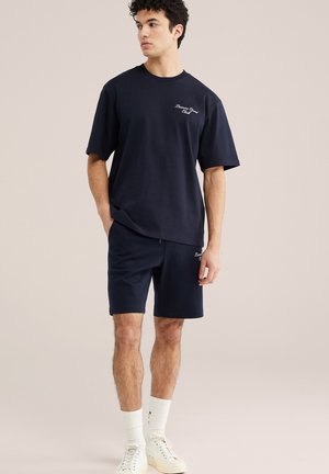 Muž oblečený v námornícky modrom neformálnom tričku a šortkách s bielymi ponožkami a teniskami, stojaci a pozerajúci sa do strany.