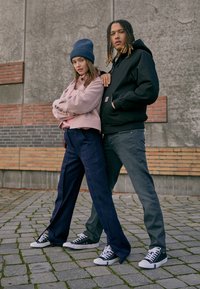 Två personer poserar framför en strukturerad grå vägg. Kvinnan har på sig en rosa kort jacka och vida blå byxor; mannen har en svart huvtröja och grå byxor. Båda bär canvas sneakers.