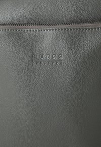 Borsa in pelle grigia testurizzata con chiusura a zip, logo "GUESS" impresso e dettagli di cucitura sottili lungo la parte superiore.