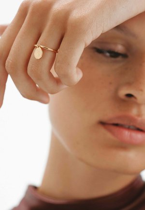 Primer plano de una persona con un anillo de oro que tiene un pequeño charm ovalado, sostenido cerca de su rostro con los ojos suavemente cerrados.