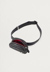 Czarna skórzana torba na pas z teksturowanym wykończeniem, czerwoną podszewką, podwójnymi zamkami błyskawicznymi i akcentami ze srebrnego metalu. Kompaktowy prostokątny kształt.