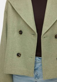 Veste courte verte à double boutonnage en mélange de laine douce, dotée de grands boutons foncés et de revers crantés, portée avec un haut foncé en dessous.