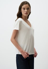 Biała koszulka typu V-neck wykonana z miękkiego materiału, z krótkimi rękawami i luźnym krojem, w połączeniu z ciemnoniebieskimi dżinsami. Gładka tekstura.