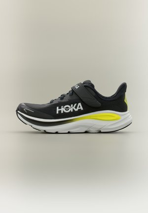 Zwart-witte Hoka hardloopschoen met gele accenten, een klittenbandsluiting en een dikke gewatteerde zool op een neutrale achtergrond.