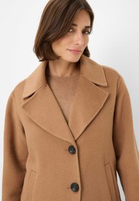 Manteau en mélange de laine camel avec un col à revers, trois boutons noirs et des poches latérales. Tissu texturé et design ajusté.