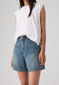 Nainen, jolla on yläosana ilman hihoja oleva valkoinen paita, joka on piilotettu korkeavyötäröisiin sinisiin farkkushortseihin mustalla vyöllä, seisoo yksinkertaista taustaa vasten.