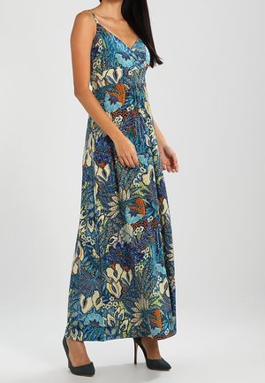 Vrouw draagt een mouwloze maxi-jurk met V-hals, bedrukt met blauwe, groene en oranje bloemen- en blaadjespatronen, gecombineerd met donkergroene pumps.
