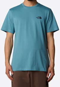 Camiseta de cuello redondo en color azul verdoso, hecha de una tela suave. Presenta mangas cortas y un pequeño logo negro en la parte izquierda del pecho. Dobladillo sin costuras.