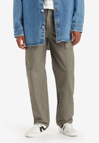 Olijfgroene cargobroek met zijzakken, gecombineerd met een lichtblauwe spijkerjas en witte sneakers met zwarte accenten.