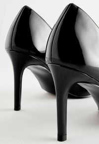 Musta lakattu nahka korkokengät, joissa on terävä kärki ja hoikka stilettokorko. Kengissä on sileä, kiiltävä pinta.