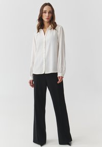 Blusa blanca de botón con detalles de encaje en los hombros, mangas largas con puños, combinada con pantalones de pierna ancha negros. Tejido suave y fluido.