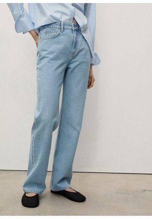Person iført lyseblå straight-leg jeans, lyseblå skjorte med knapper og sorte vævede flade sko, stående mod en ensartet lys væg.