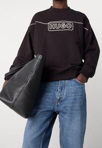 Sudadera negra con el logotipo blanco "HUGO", cuello redondo acanalado y hombros caídos, combinada con jeans de mezclilla azul claro y un bolso de cuero negro.