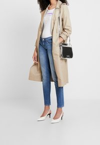 Mujer vestida con abrigo trinchera beige, camiseta gráfica blanca, jeans azules, tacones de kitten blancos y llevando un bolso negro y plateado de Calvin Klein.